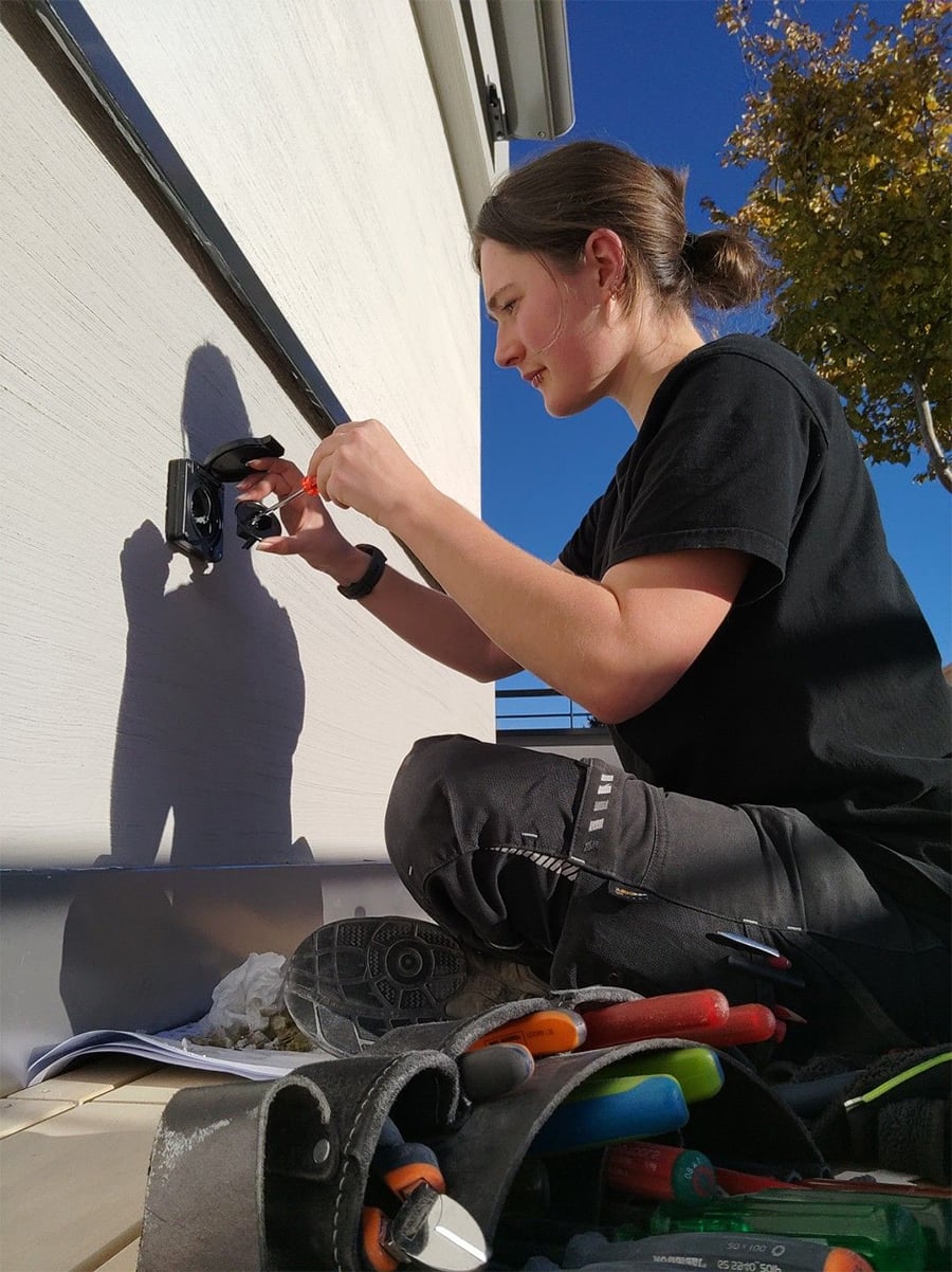 A person kneels outside, installing or repairing an electrical outlet on a building wall. Tools and a tool belt are visible in the foreground, and a tree with autumn leaves is in the background.