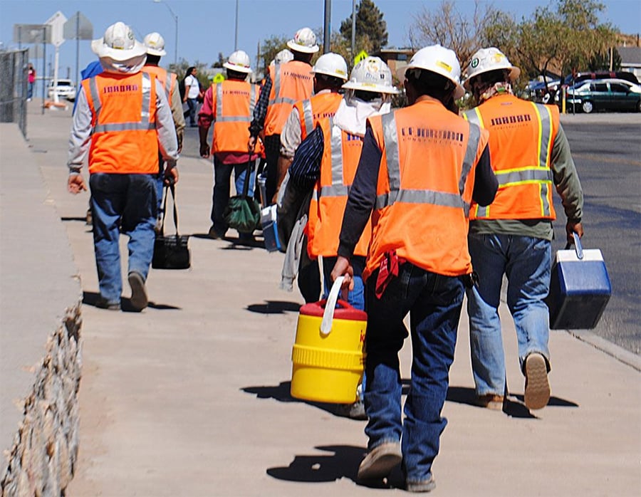 A group of construction workers wearing orange safety vests and helmets walk down a sidewalk, carrying lunchboxes and coolers.