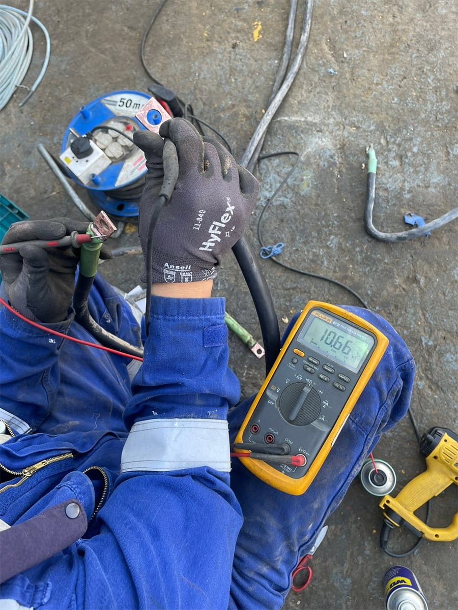 A person in blue workwear and gloves uses a digital multimeter to measure voltage on a black cable, with various tools and cables scattered on the ground nearby.
