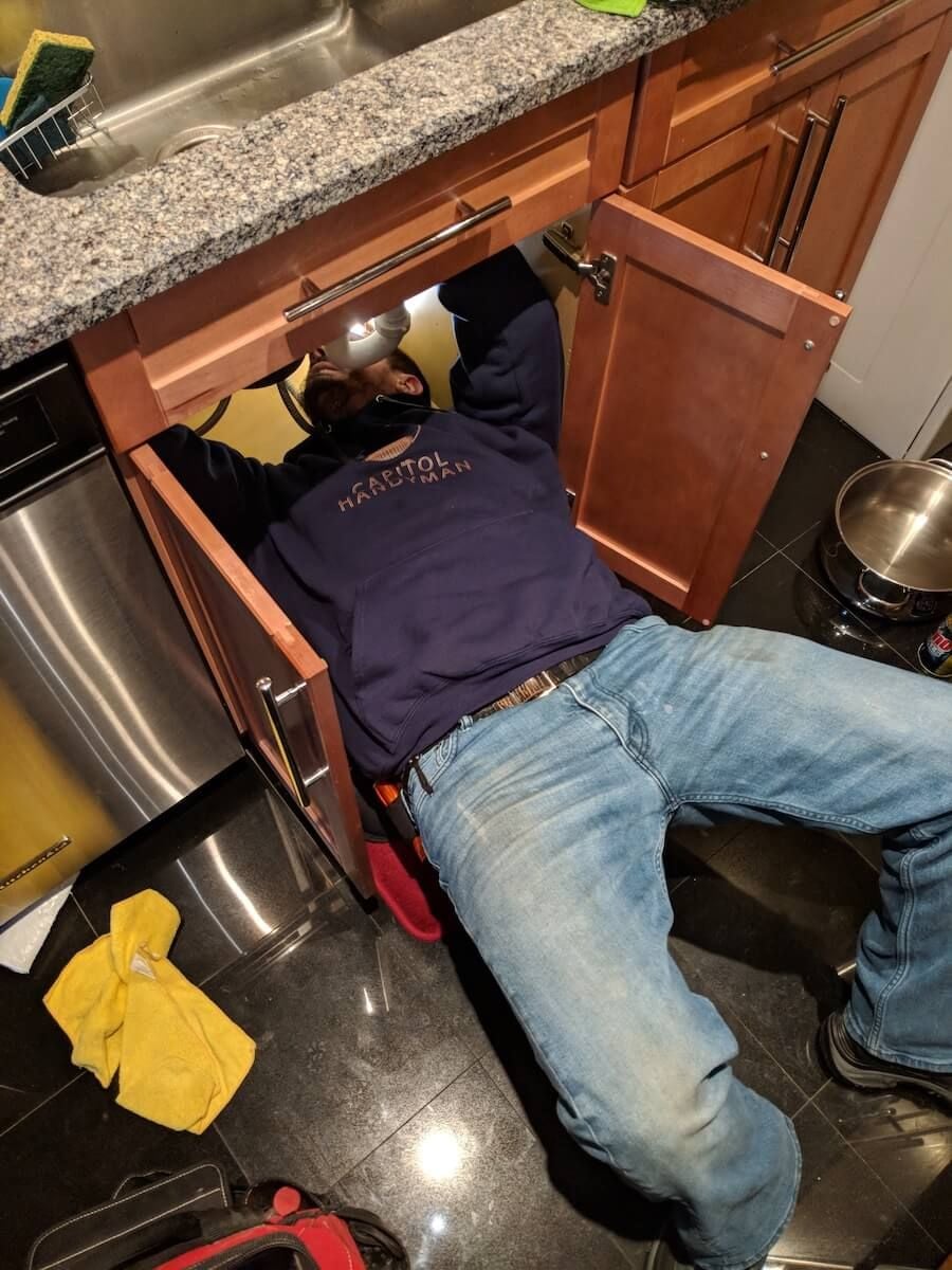 A person wearing jeans and a hoodie is lying on their back under a kitchen sink, working with tools. Cabinet doors are open, and cleaning supplies are nearby on the floor.