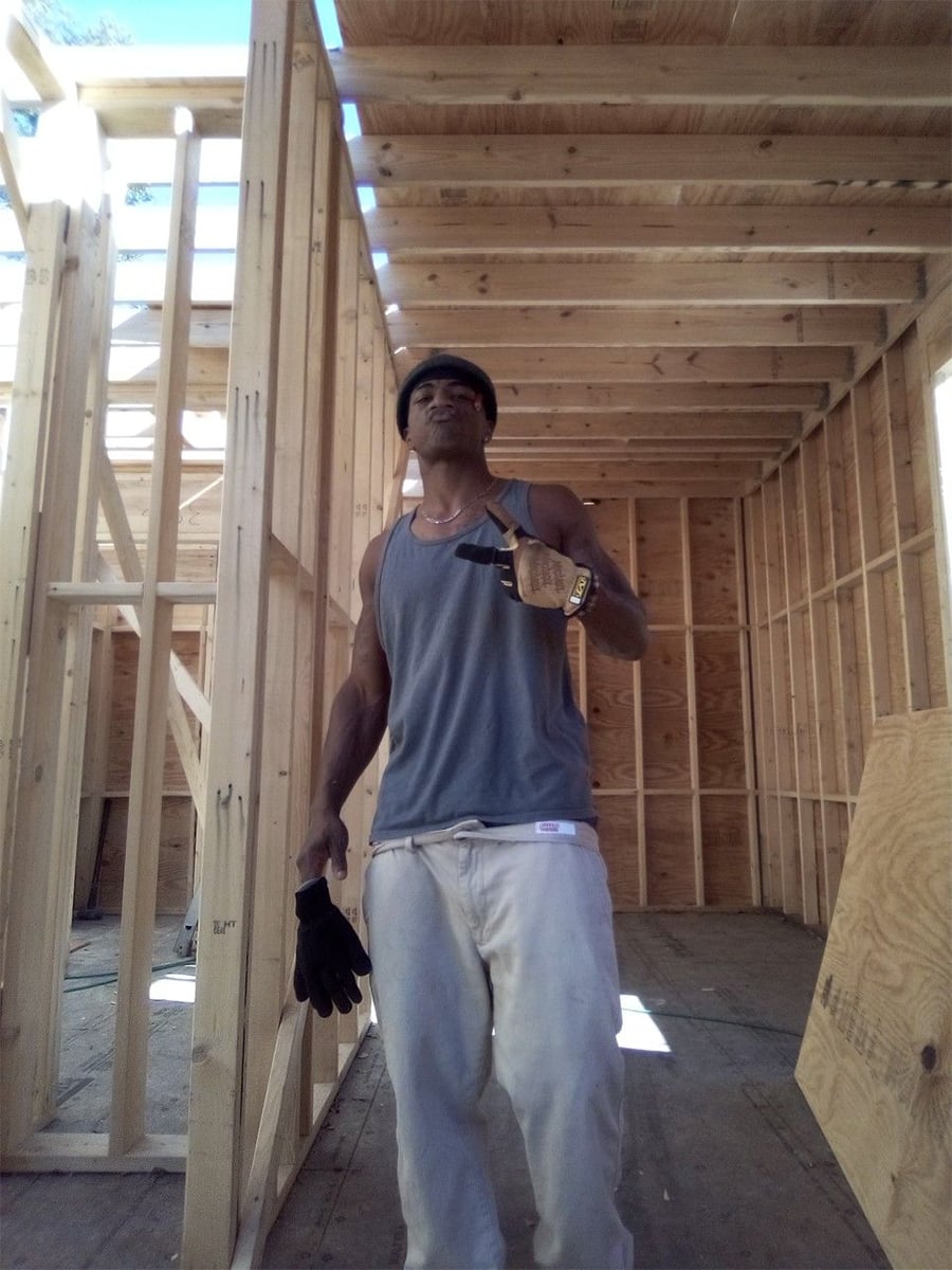 A man in a gray tank top and white pants stands inside a wooden building under construction, holding gloves and gesturing with his hand.