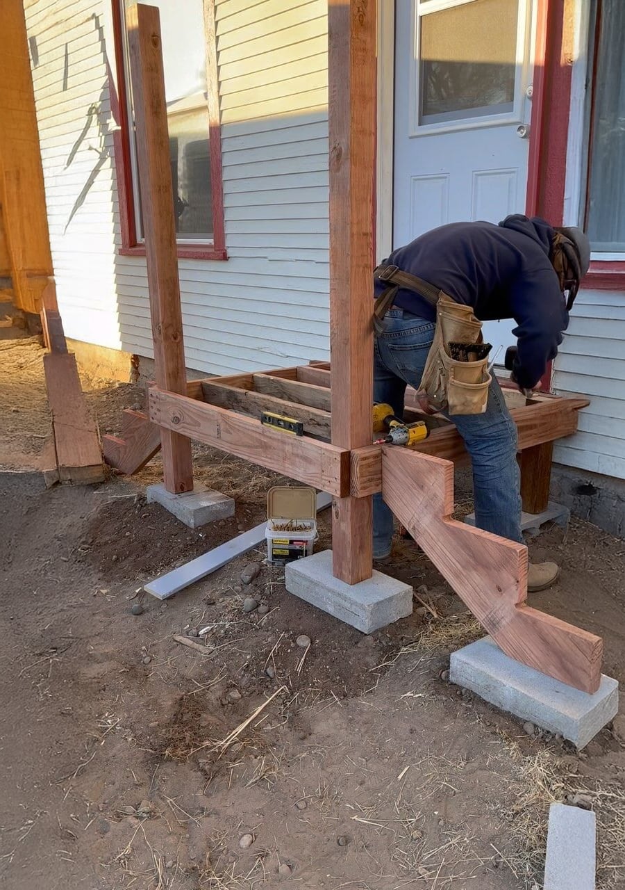 A person wearing a tool belt is building a wooden porch with four upright posts on concrete blocks in front of a white house with red trim.