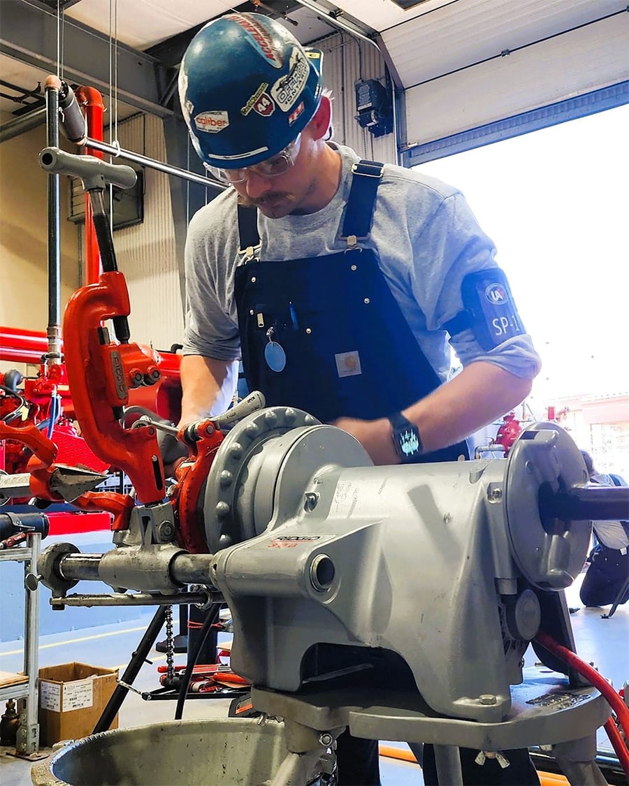 A worker in safety gear uses a pipe threading machine indoors, focusing on the task. Tools and pipes surround the workspace.
