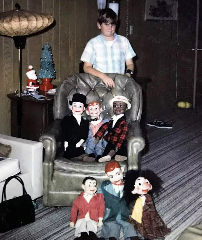A boy stands behind a chair displaying six ventriloquist dummies in a living room with wood-paneled walls, a lamp, and holiday decorations.