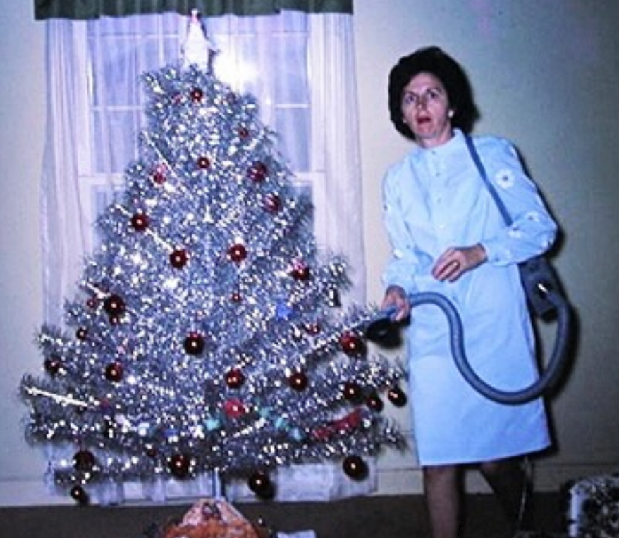A woman in a light blue dress stands by a silver tinsel Christmas tree decorated with red ornaments, holding a vacuum cleaner hose in a living room.