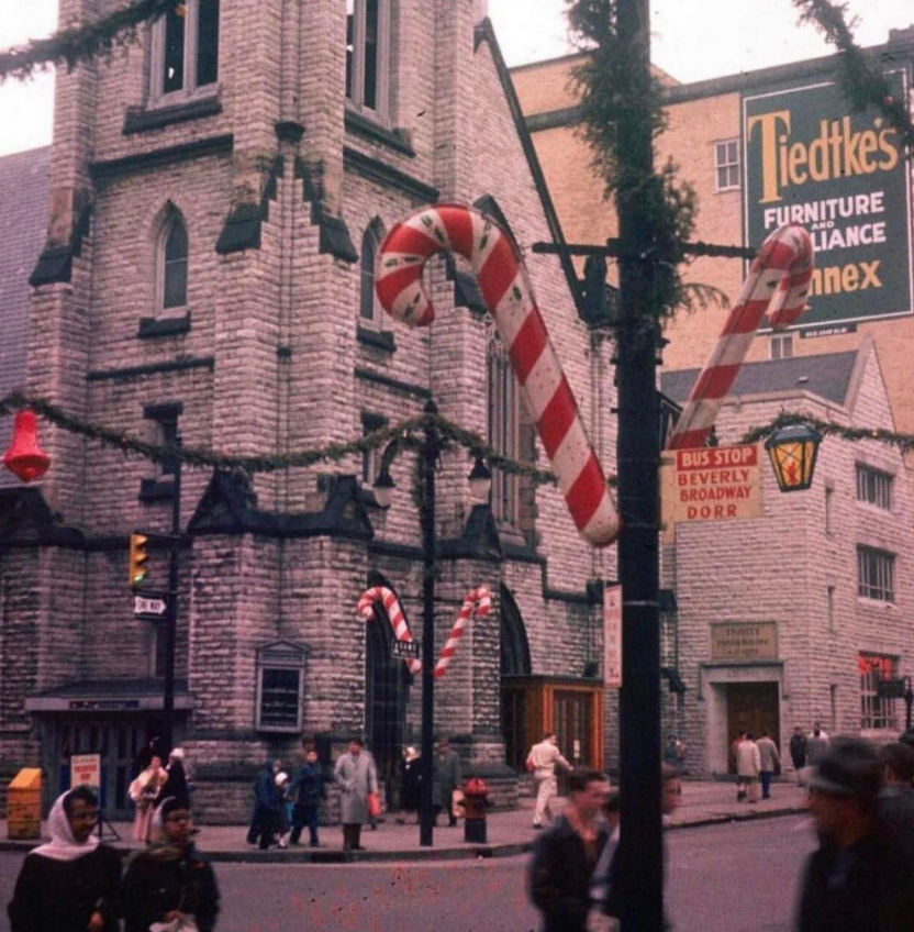 People walk past a stone church decorated with large candy canes and garland. Signs for "Tiedtke's Furniture Annex" and a bus stop are visible. The scene appears festive and busy.