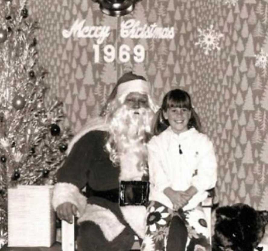 A young girl sits on Santa’s lap beside a decorated Christmas tree, with “Merry Christmas 1969” and snowflakes on the patterned wall behind them. The scene is in black and white.