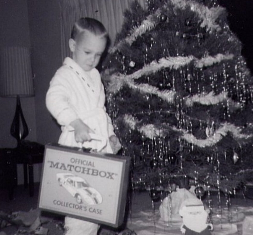 A young child in pajamas holds an "Official Matchbox Collector's Case," standing near a Christmas tree decorated with tinsel and ornaments. Wrapped presents and a Santa decoration are on the floor. The photo is black and white.
