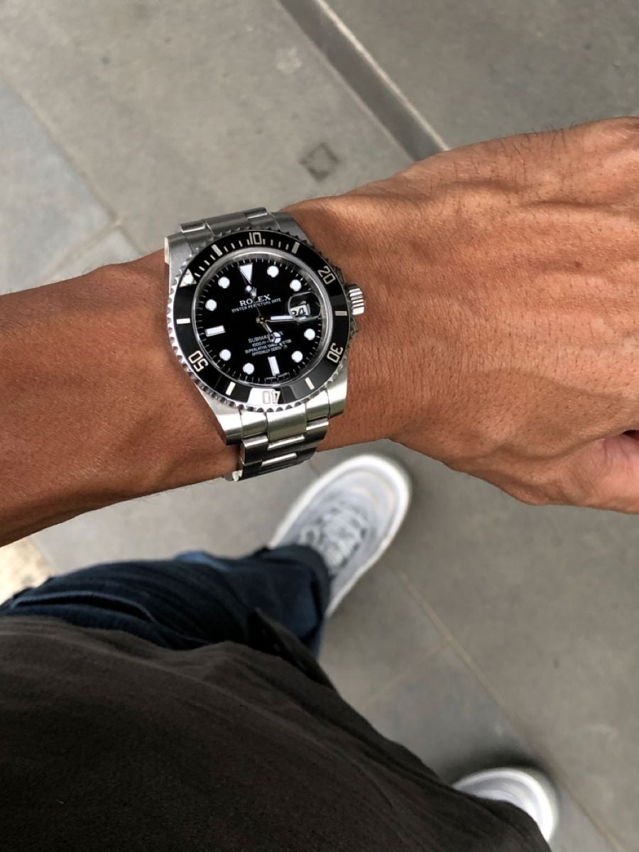 A person wearing a silver wristwatch with a black face and white details, standing on a gray pavement. Only their hand, watch, part of their arm, jeans, and white sneakers are visible.