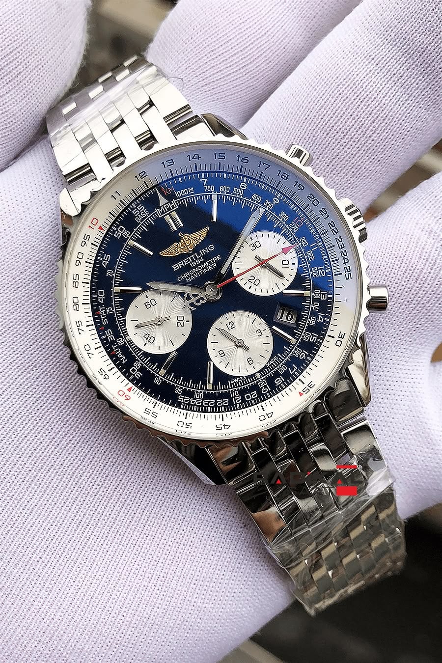 A gloved hand holds a luxury Breitling chronograph watch with a blue dial, three subdials, and a metal bracelet. The watch face displays various markings and the brand logo.