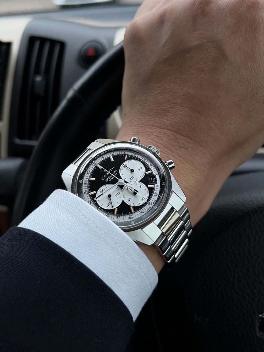 A close-up of a person’s wrist wearing a silver chronograph wristwatch with a black and white dial, holding a steering wheel inside a car.