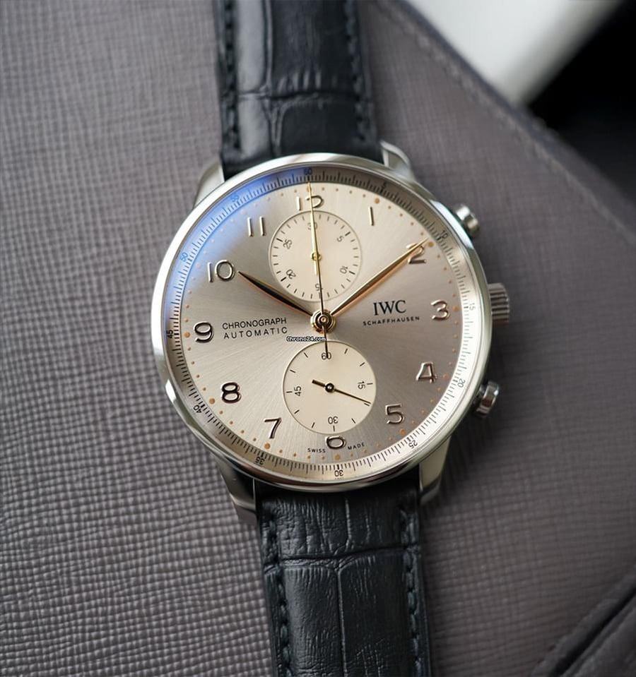 A silver IWC chronograph wristwatch with a black leather strap rests on a textured gray surface. The watch face features gold numbers, two subdials, and slim gold hands.