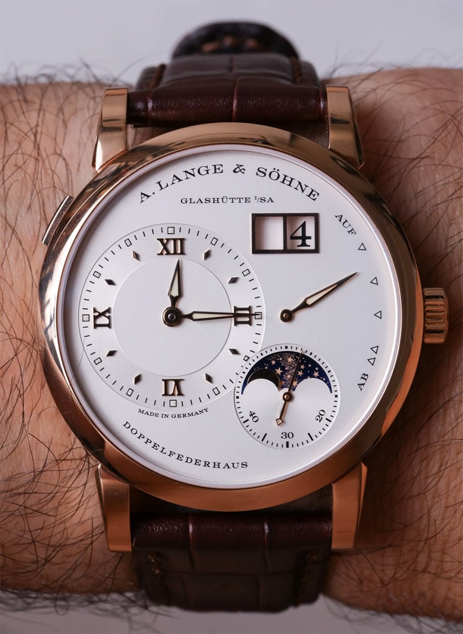 A gold A. Lange & Söhne wristwatch with a white dial, Roman numerals, moonphase subdial, date window, and brown leather strap on a person’s wrist.