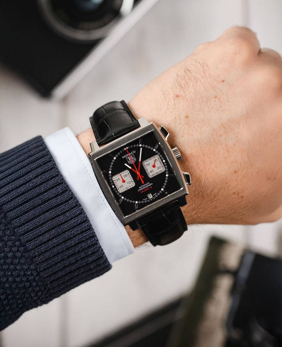 A person wears a black square chronograph wristwatch with a leather strap and red hands. The watch face has two white subdials. The person is dressed in a blue sweater with a light blue shirt underneath.