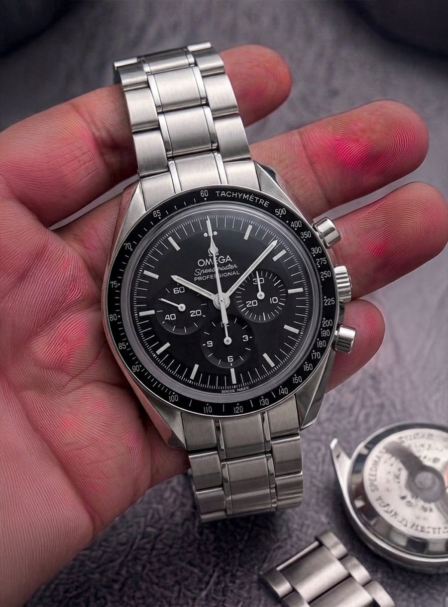 A hand holds an Omega Speedmaster wristwatch with a black dial and stainless steel bracelet. Another watch and extra bracelet links lie on a gray surface in the background.