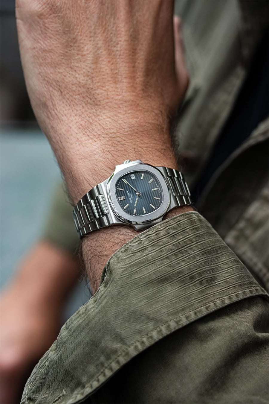 A close-up of a person’s wrist wearing a silver luxury wristwatch with a dark face and metal band. The person is dressed in a green jacket.