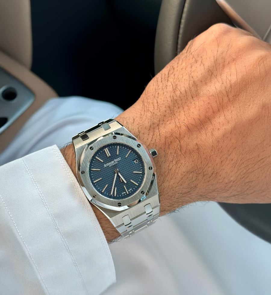 A close-up of a person’s wrist wearing a silver Audemars Piguet watch with a blue face, holding a steering wheel.