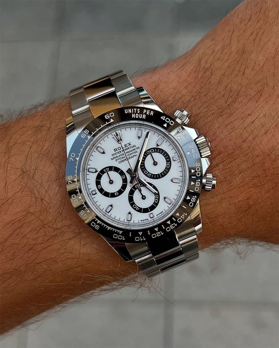 A close-up of a stainless steel Rolex Daytona watch with a white dial and black subdials, worn on a person's wrist against a blurred gray background.