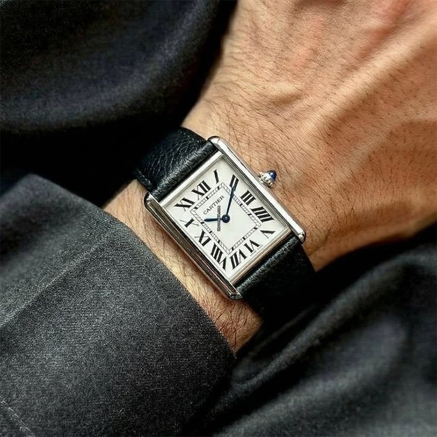 A close-up of a wrist wearing a rectangular Cartier watch with a white dial, black Roman numerals, blue hands, and a black leather strap. The person is dressed in a dark gray shirt.