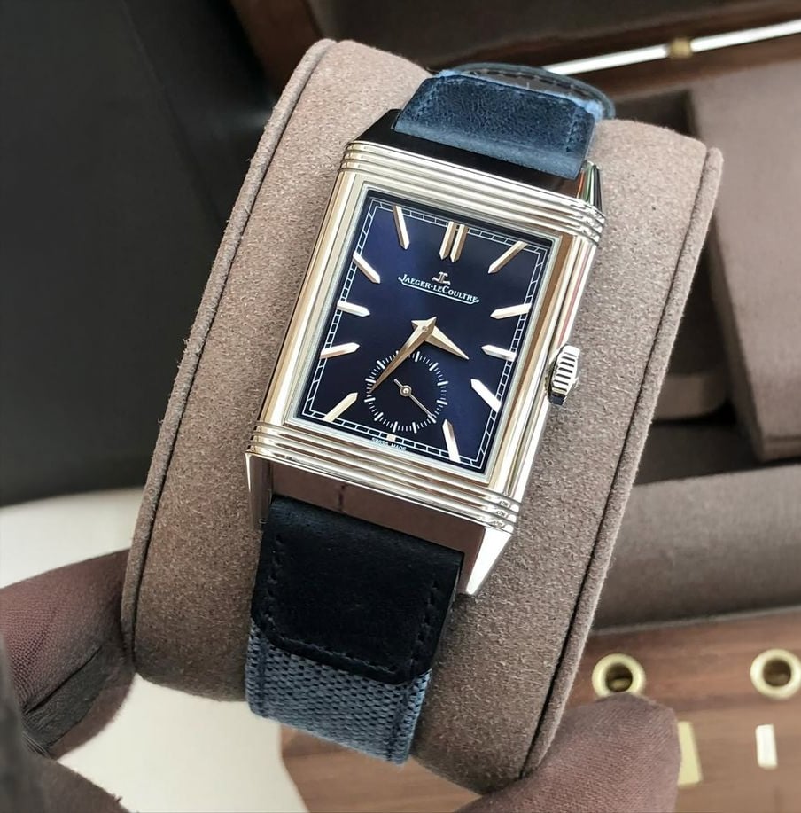 A rectangular Jaeger-LeCoultre wristwatch with a blue dial, silver hour markers, and a blue leather strap, displayed on a brown watch holder.