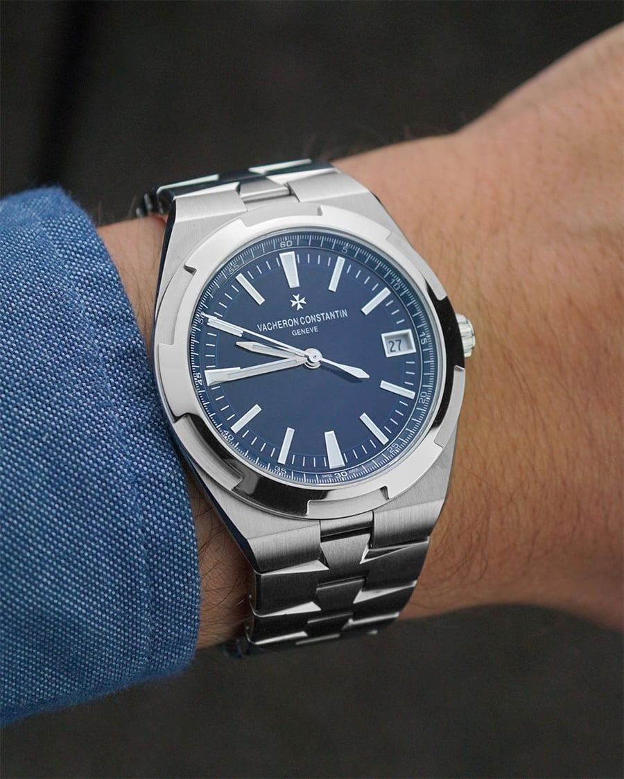 A silver Vacheron Constantin watch with a blue face and metal band is worn on a person's wrist, who is dressed in a blue long-sleeve shirt. The time on the watch is 3:16.