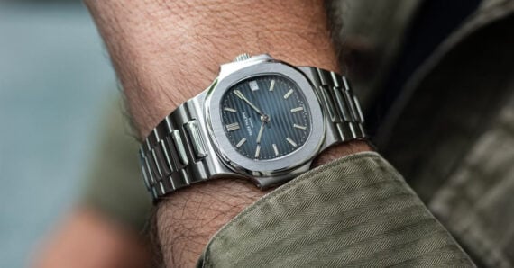 A close-up of a silver wristwatch with a dark, striped dial on a person's wrist, who is wearing a green textured jacket.