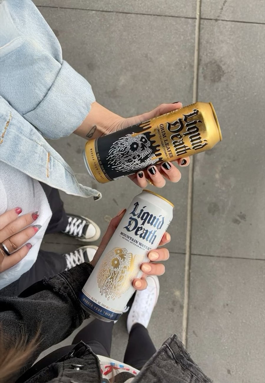 Two people holding Liquid Death mountain water cans—one black and gold, one white and gold—standing on a gray sidewalk, wearing casual clothes and sneakers.