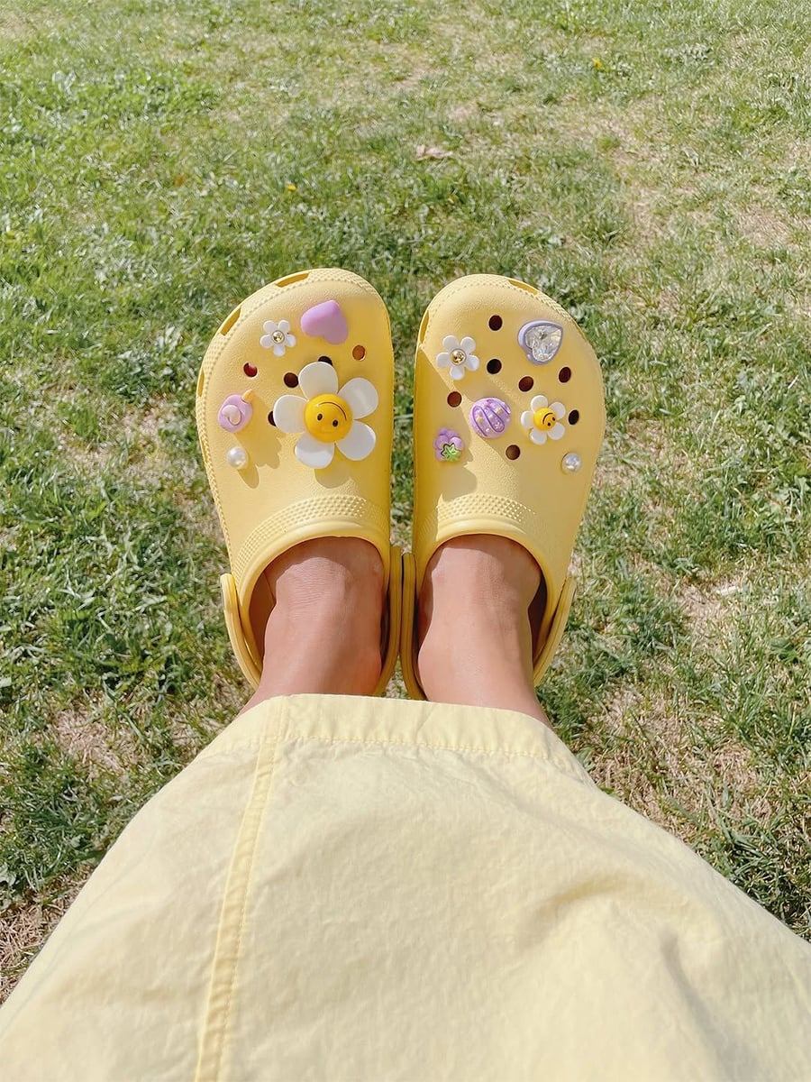 Person wearing yellow Crocs decorated with cute charms like flowers and a smiley face, sitting on grass. They are also wearing a yellow skirt or dress.