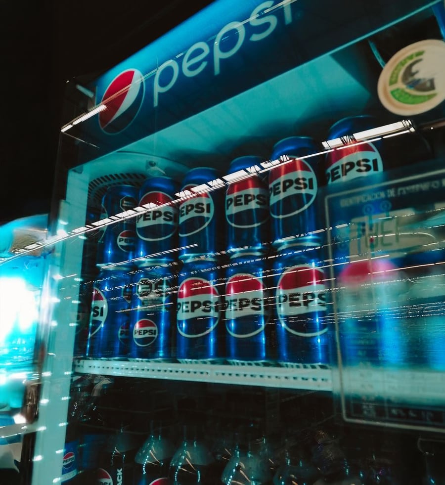 Cans of Pepsi are stacked inside a refrigerator with a Pepsi logo on top. The refrigerator door reflects light, creating a blue glow and blurred reflections. Bottles are visible on the lower shelf.