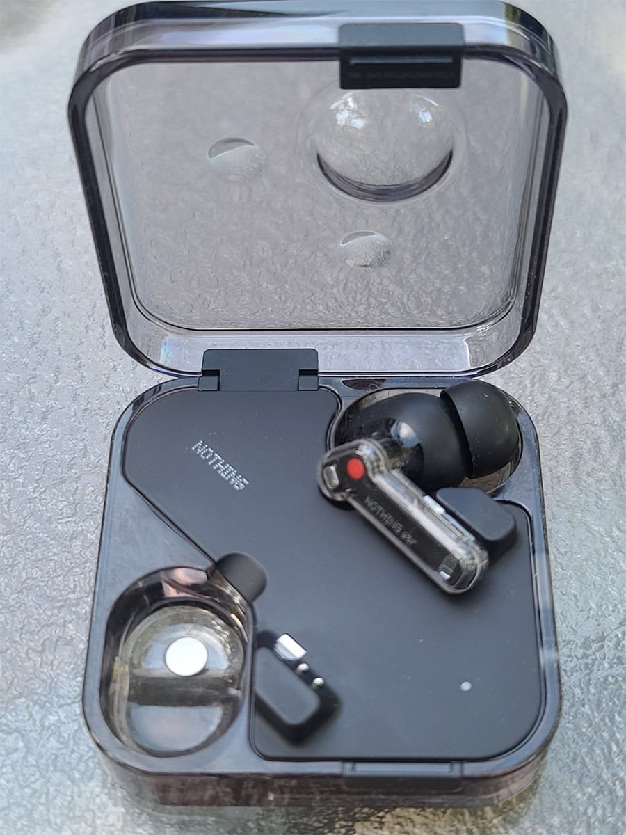 A pair of black wireless earbuds in a transparent charging case, resting on a glass surface. One earbud is placed in the case, and the other is slightly out, showing the "NOTHING" branding.