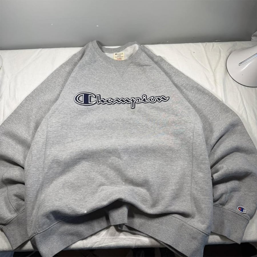A gray Champion sweatshirt with the brand’s logo in navy and white across the chest, laid flat on a white surface under soft lighting.