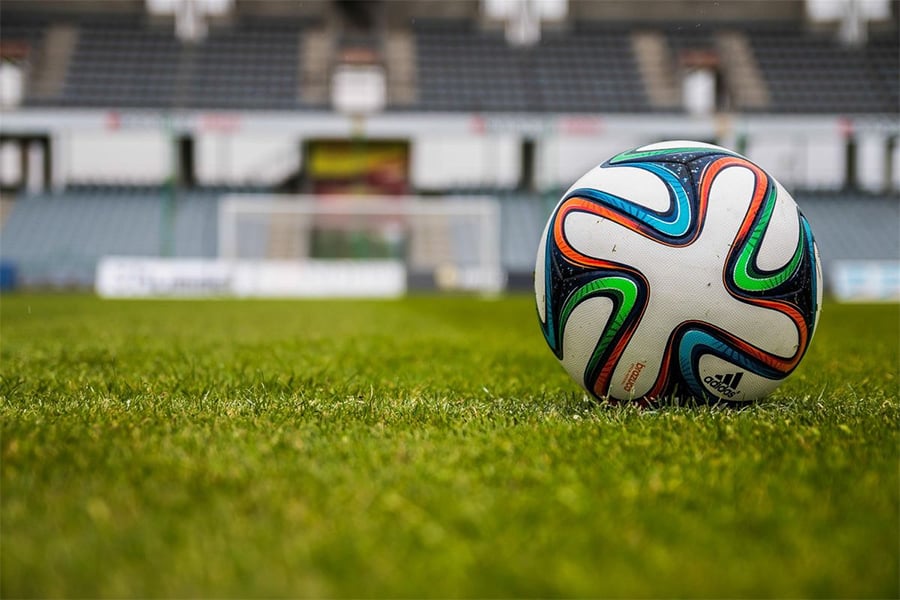 A close-up of a colorful soccer ball resting on green grass, with a blurred stadium and empty seats in the background.