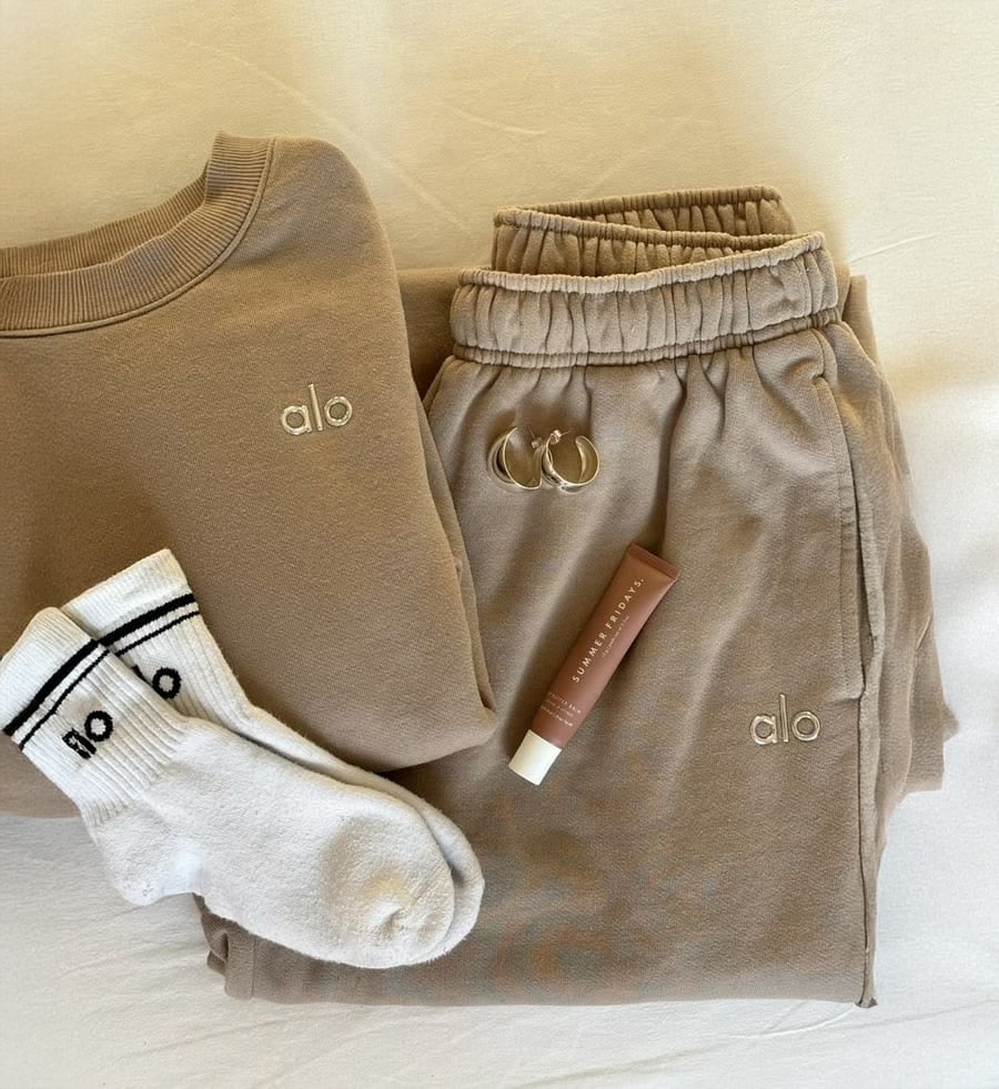 Folded beige Alo sweatshirt and sweatpants, white Alo socks, two silver hoop earrings, and a tube of tinted lip balm are neatly arranged on a cream surface.
