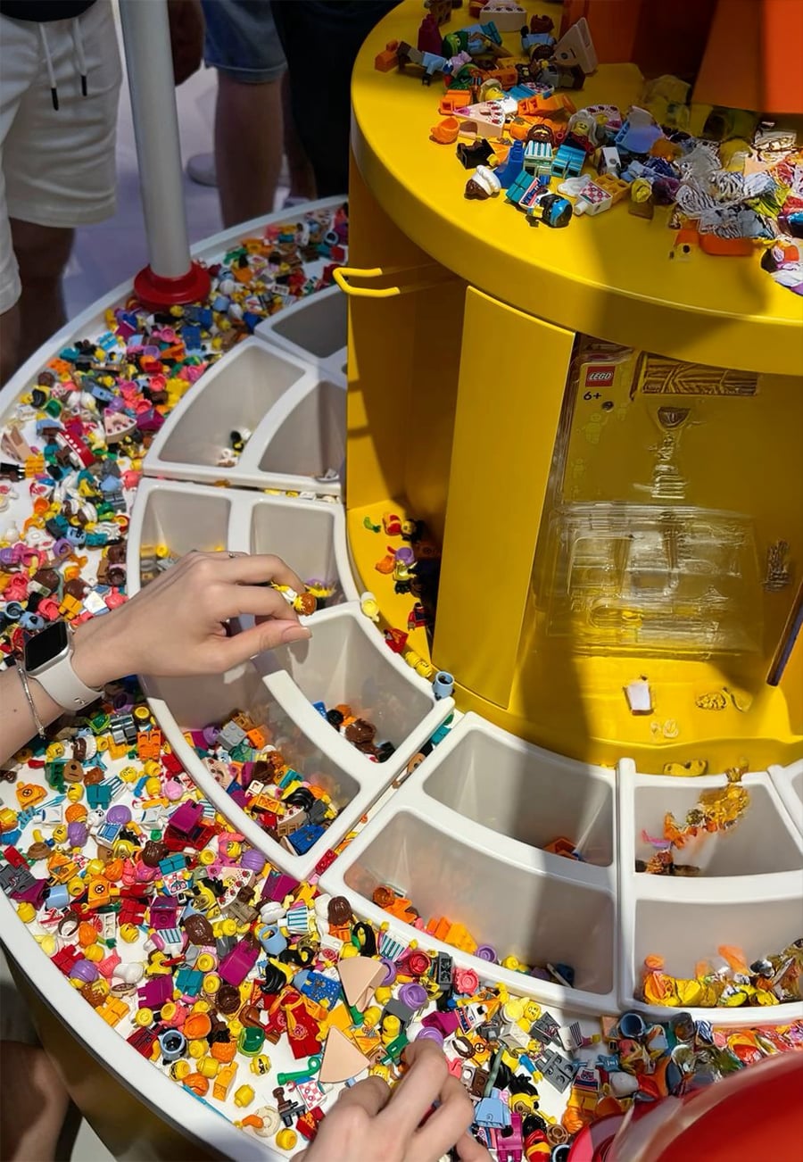 A person sorts through colorful LEGO pieces in organized bins on a round yellow table, surrounded by other people. The table has various LEGO parts, including heads, torsos, and accessories.