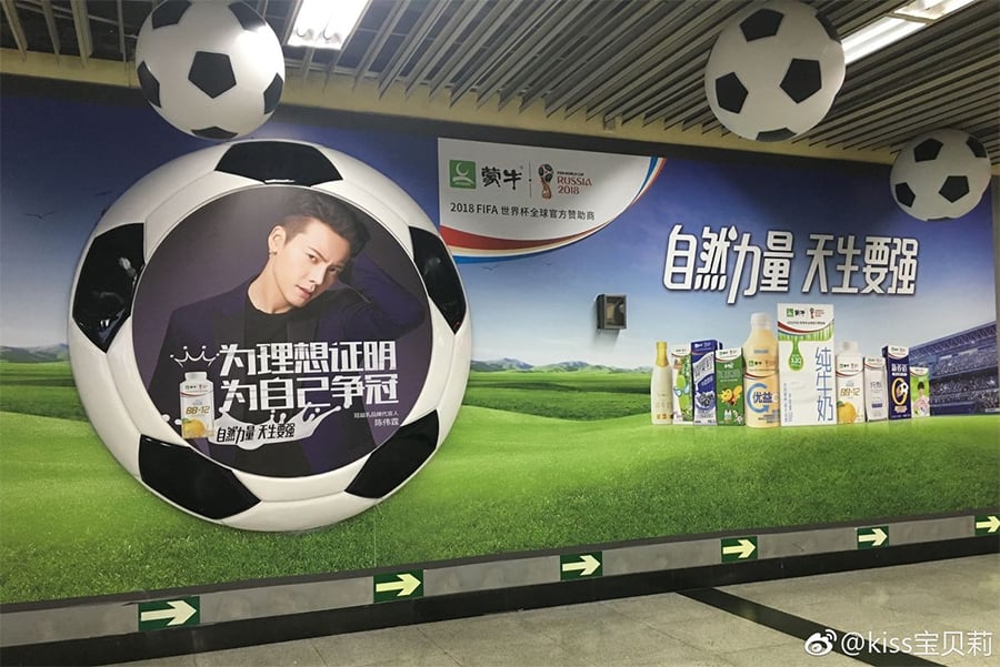 A subway station wall decorated with soccer balls, a large poster of a man, and a display of beverage products; themed for the 2018 FIFA World Cup with Chinese text and green directional arrows on the floor.