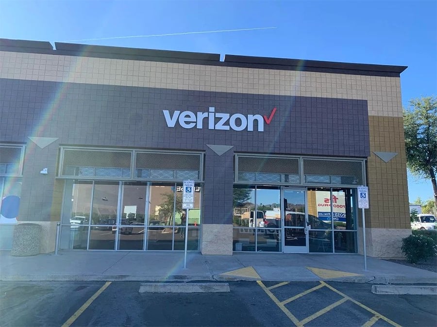 A Verizon store exterior with large windows, a Verizon sign above the entrance, and parking spots with accessibility signs in front. Sunlight creates lens flares across the image.