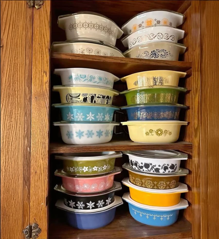 A wooden cabinet shelf filled with stacks of colorful, vintage Pyrex dishes, each with unique patterns and designs, organized by size and shape.
