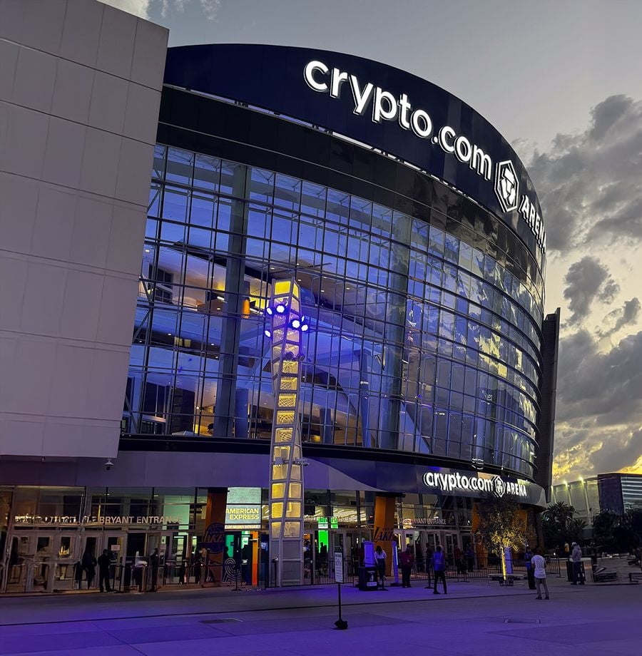Glass exterior of Crypto.com Arena at dusk, with illuminated signs and people walking near the entrance under a cloudy sky.