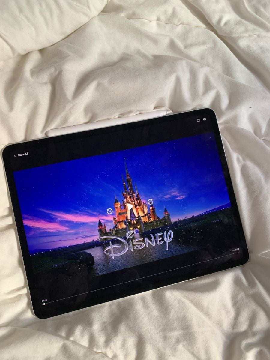 A tablet on a white bed displays the Disney castle logo on its screen, indicating a Disney movie is about to start.