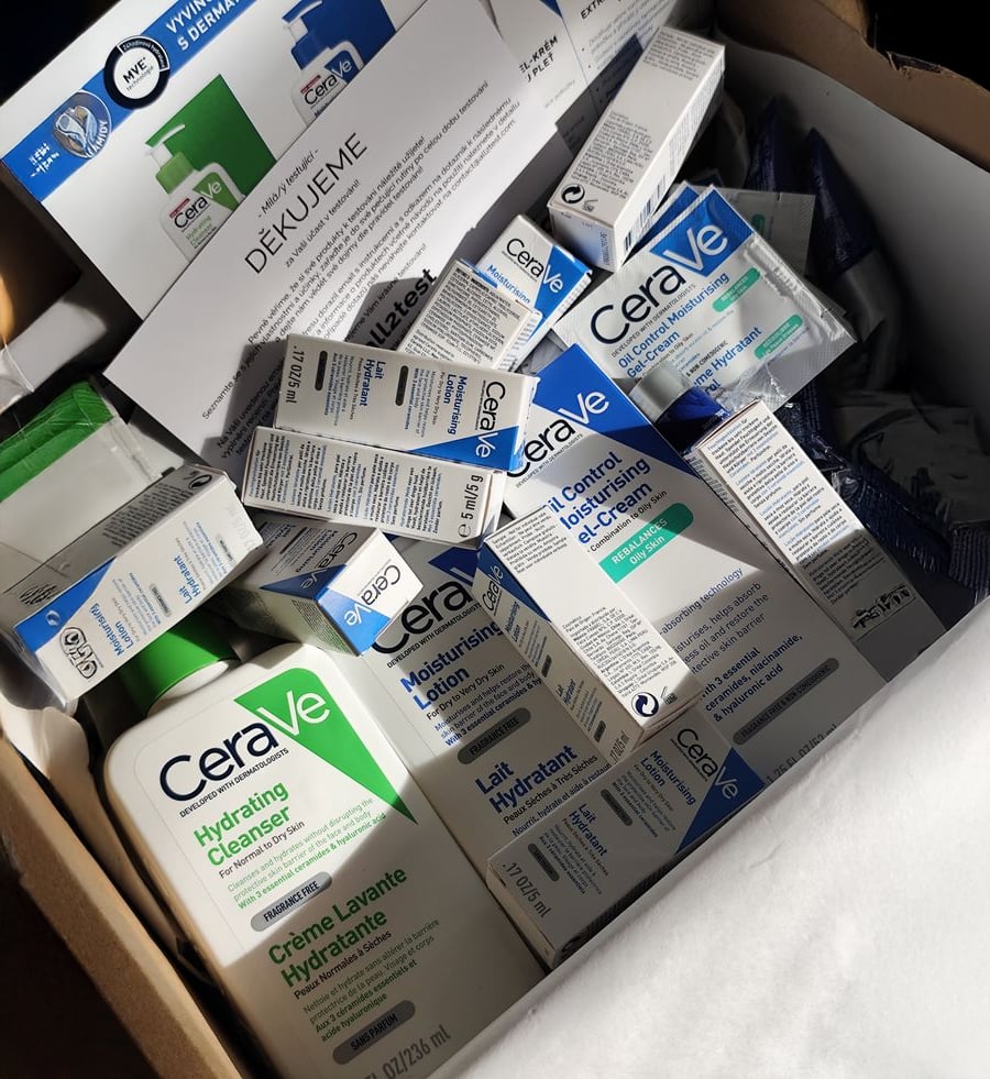 A box filled with various CeraVe skincare products, including Hydrating Cleanser bottles, creams, and lotions, all in white and blue packaging. Sunlight partially illuminates the items.