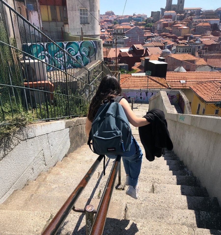 A person with long dark hair and a backpack walks down steep stone stairs overlooking a city with red-tiled roofs and graffiti on nearby walls under a clear sky.