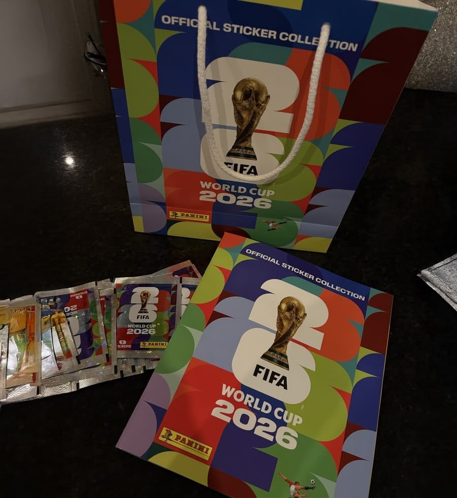 FIFA World Cup 2026 sticker album, gift bag, and Panini sticker packs are displayed on a dark surface, all featuring colorful, official tournament branding.