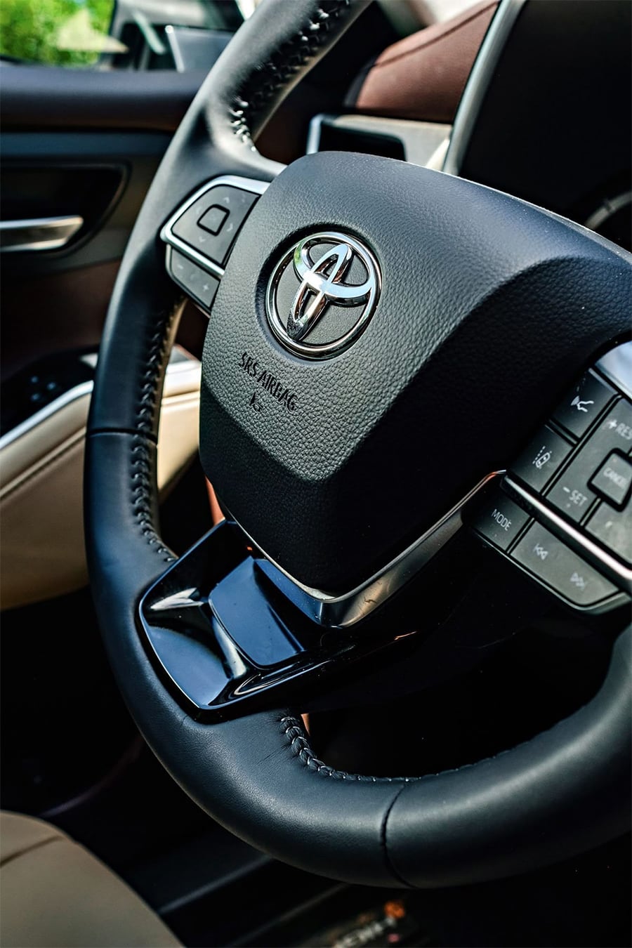 Close-up of a Toyota car steering wheel with visible control buttons, black leather finish, and part of the dashboard and interior in the background.