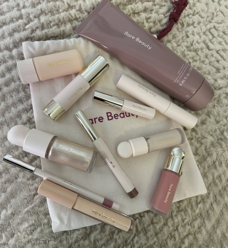 A collection of Rare Beauty makeup products arranged on a textured beige surface and a branded drawstring pouch, including tubes, bottles, and sticks in soft, neutral tones.