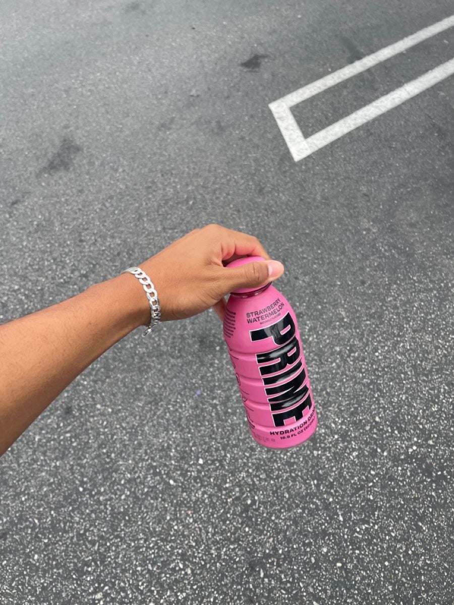 A hand holding a pink bottle of PRIME hydration drink labeled "Strawberry Watermelon" over an asphalt parking lot with white lines.