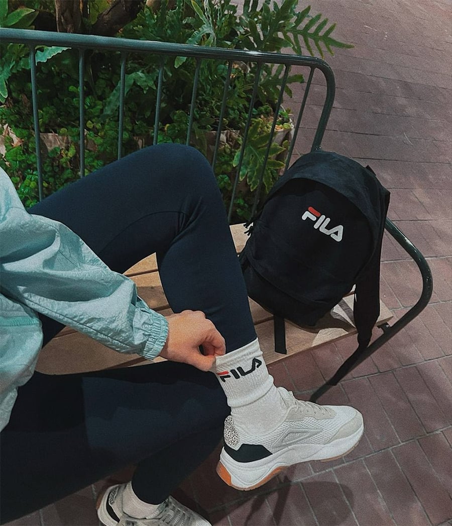 Person in athletic wear sits on a bench with one leg crossed, wearing white FILA shoes and socks. A black FILA backpack rests nearby. Green plants and tiled pavement are visible in the background.