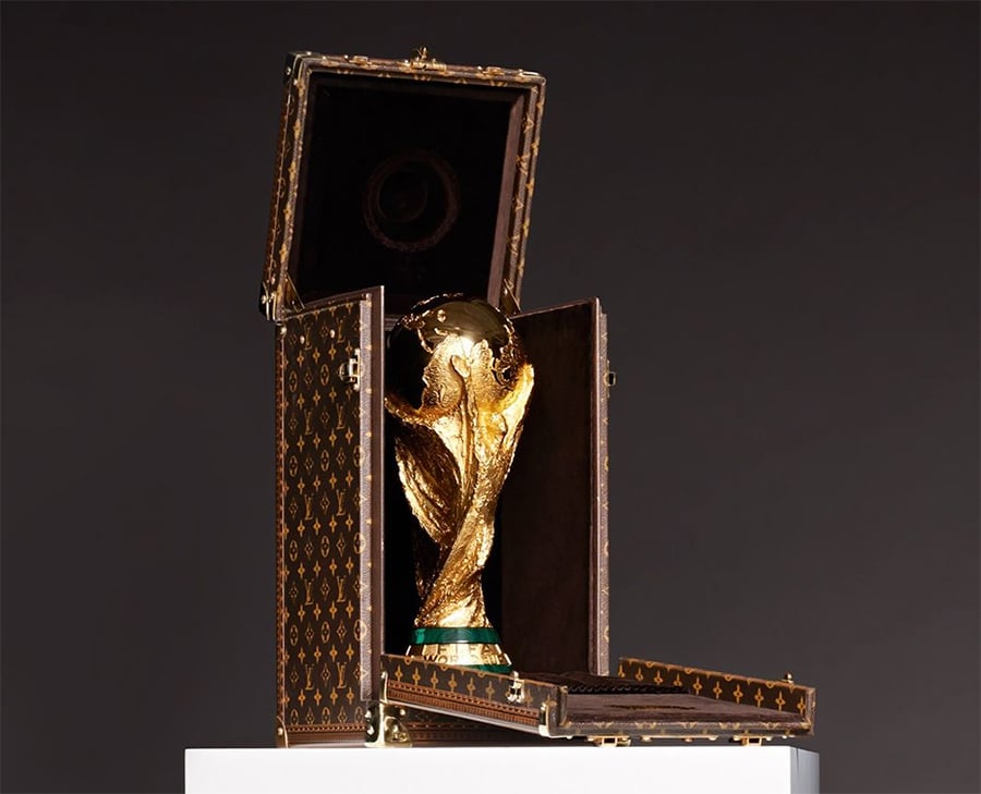 A gold trophy with green accents displayed in an open, upright, brown Louis Vuitton case with gold clasps, sitting on a white pedestal against a dark background.