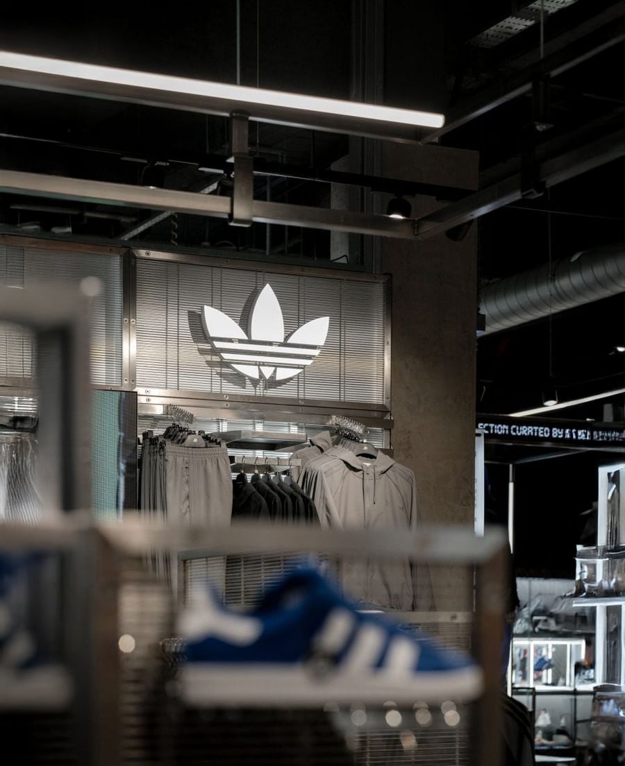 Adidas store interior with clothes hanging under a large illuminated Adidas trefoil logo; in the foreground, a blurred blue and white Adidas shoe is visible.