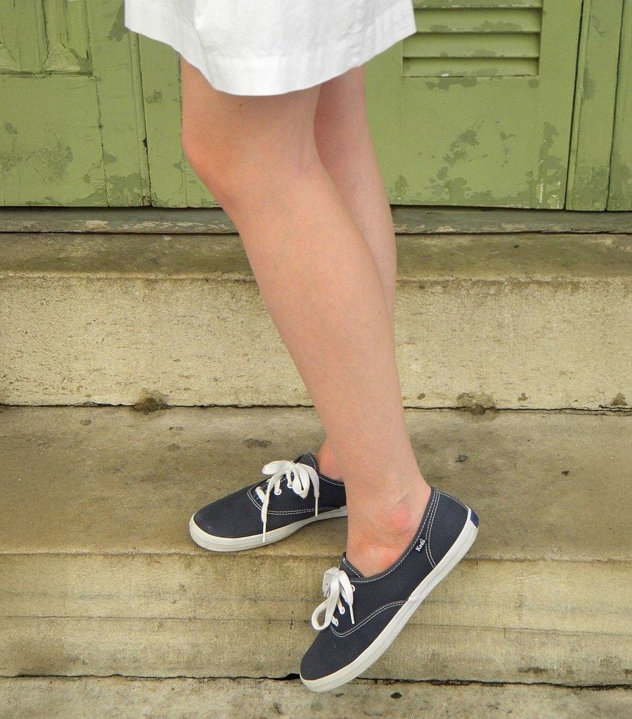 A person wearing a white skirt and navy blue sneakers with white laces stands on concrete steps in front of a green door.