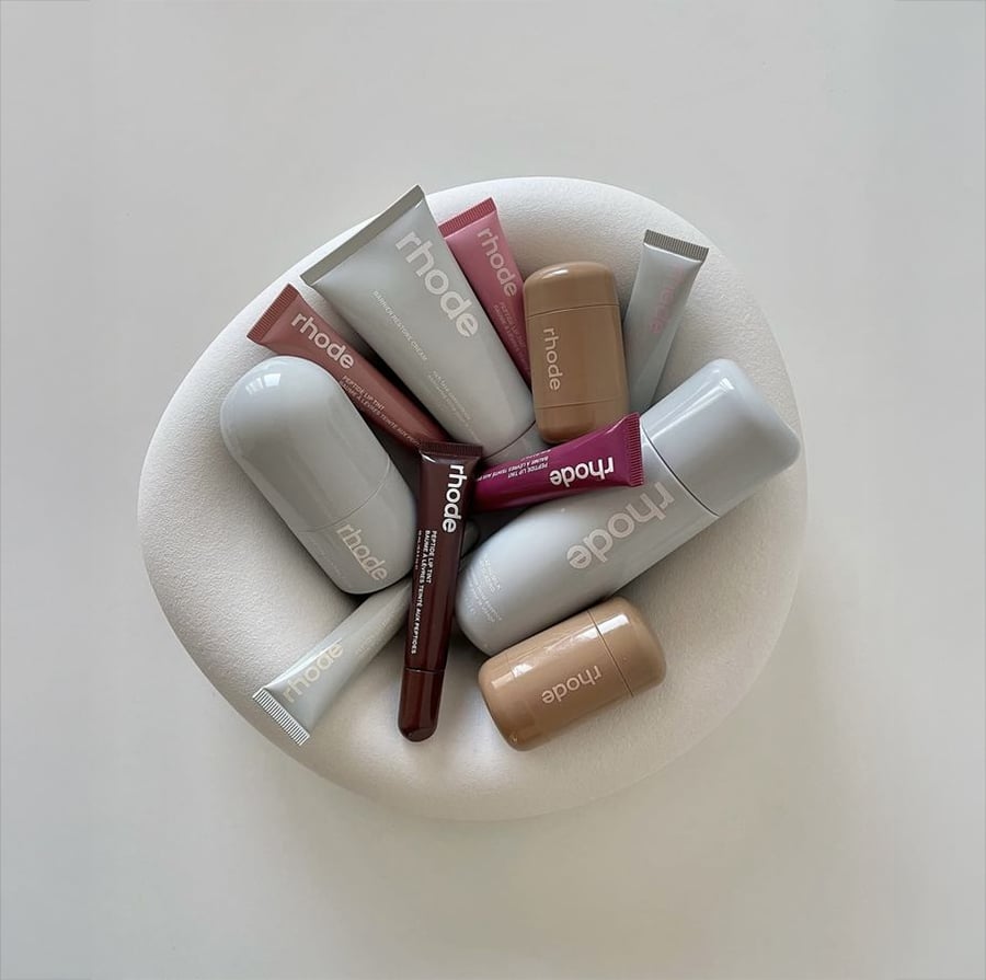 A variety of rhode skincare and beauty products are arranged in a circle on a white textured pad, including tubes, bottles, and sticks in neutral and pinkish tones.
